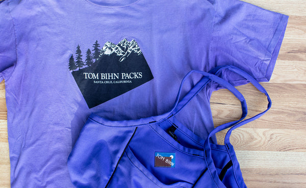A TOM BIHN Logo t-shirt from the 1990's, the Santa Cruz days; next to it is an Organic Cotton Truck in the color Santa Cruz purple. The t-shirt inspired the color for the tote.